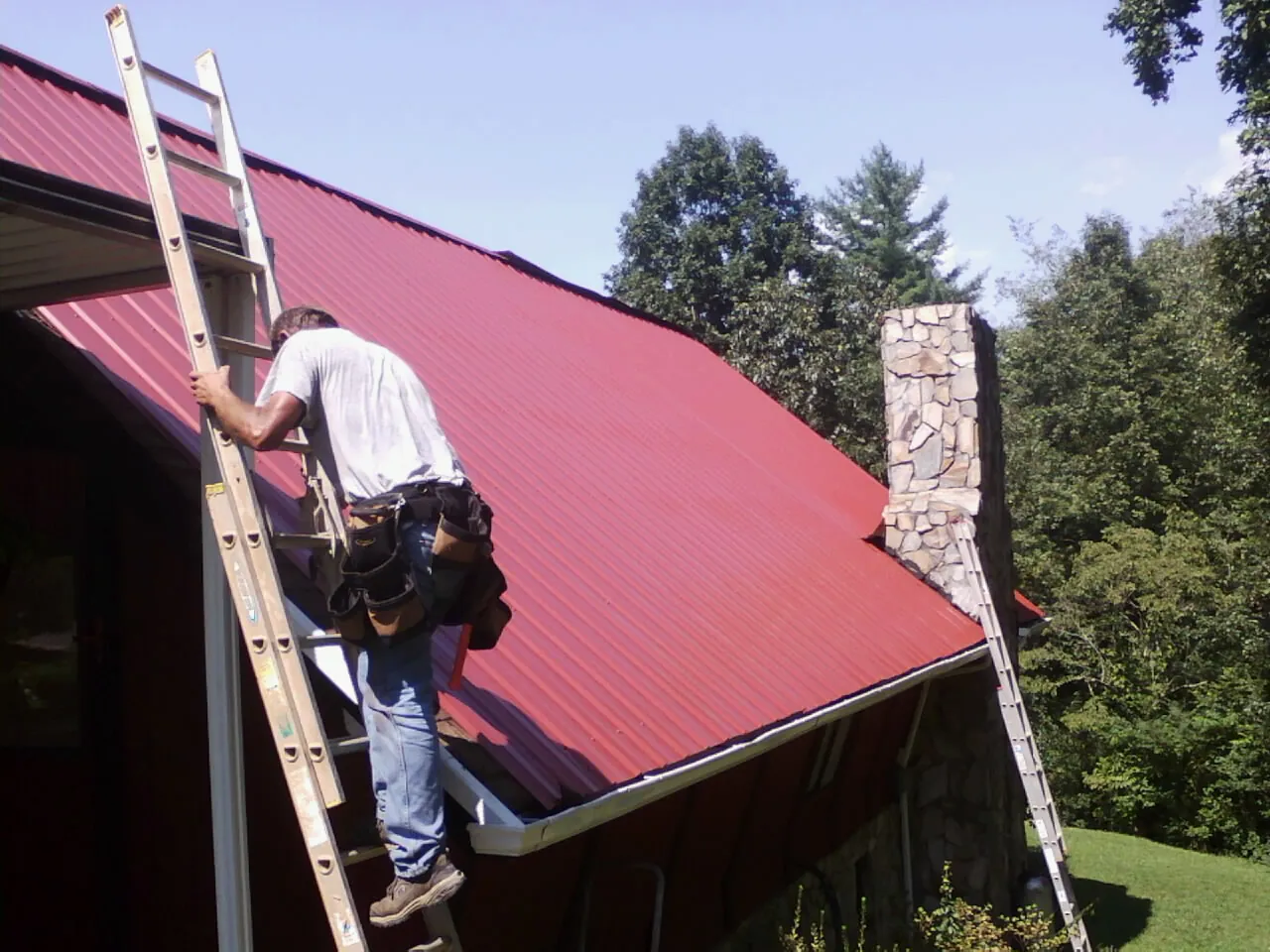 Roofer on a ladder beside a red metal roof and stone chimney — Soffit & Fascia Repair by Roofing Brothers in East Pembroke